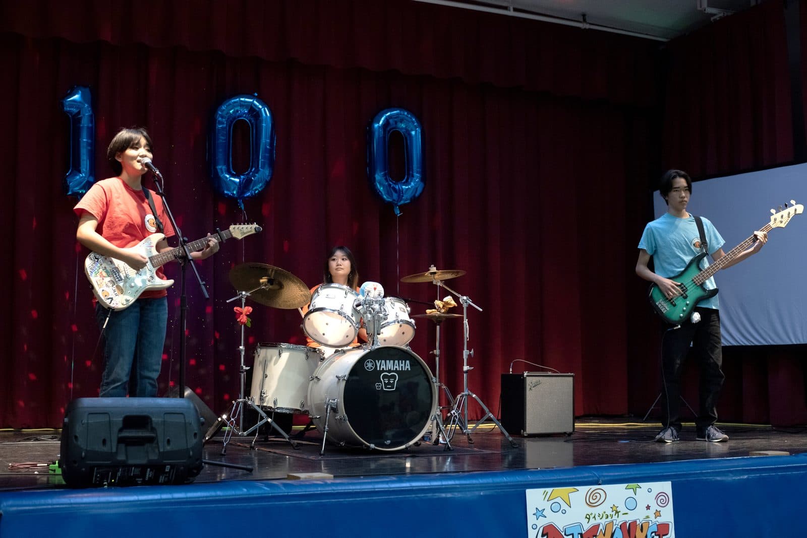 A band playing on stage beneath big blue “100” balloons