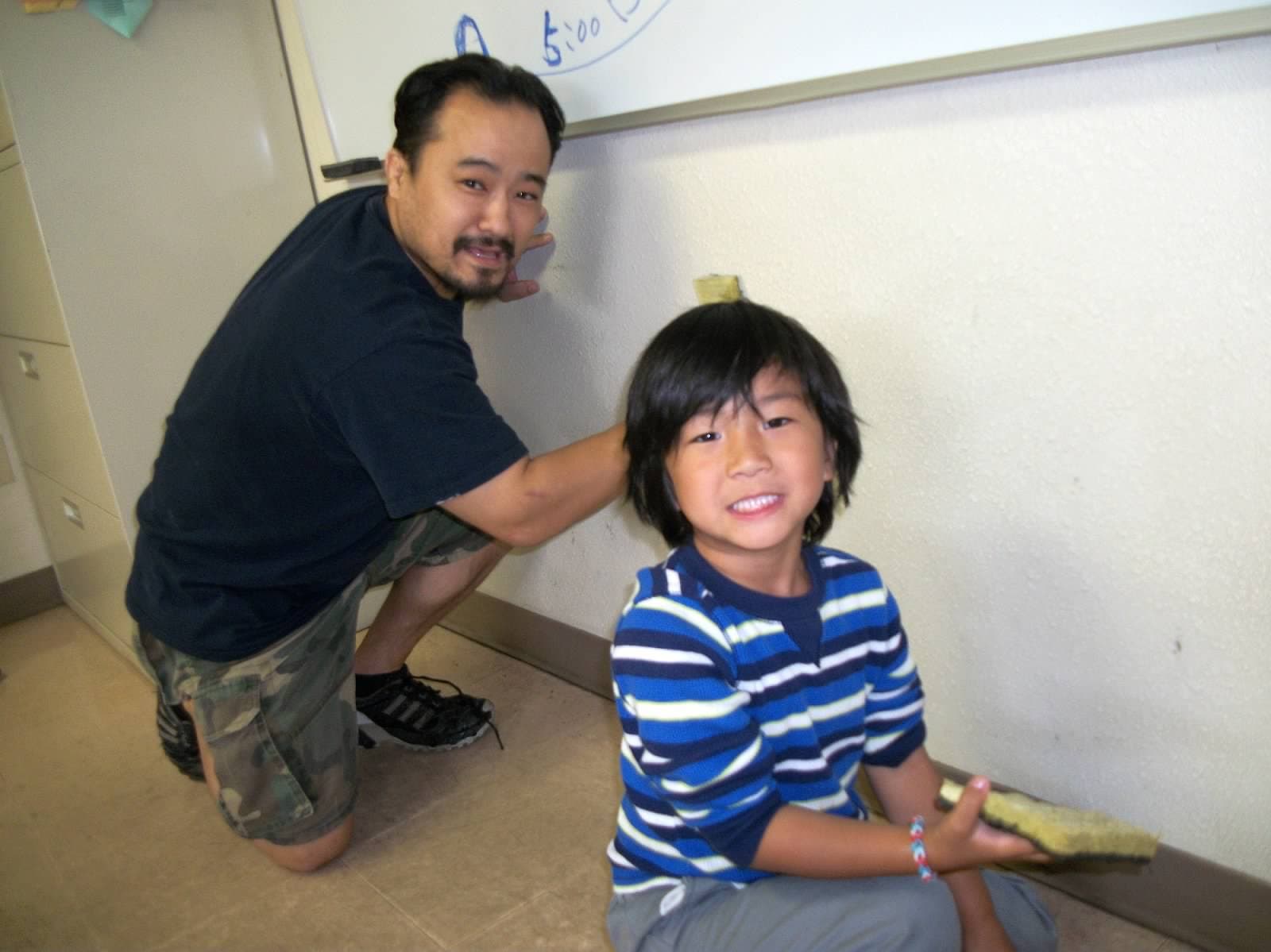 A father and his son scrubbing a classroom wall on community clean-up day