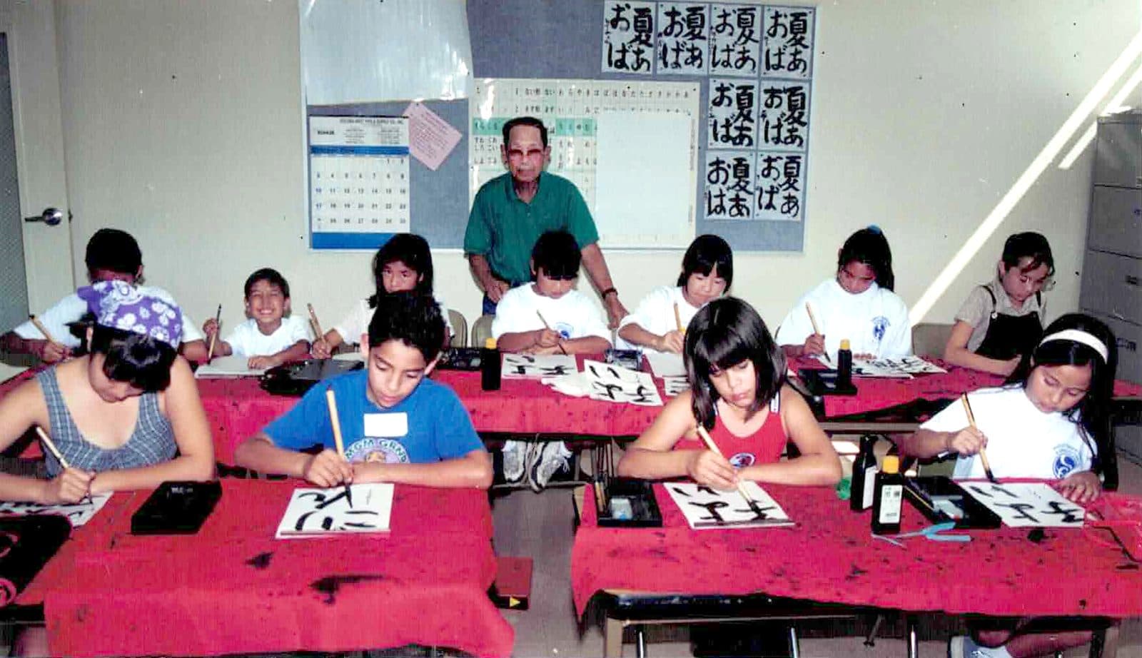 Students practicing brush calligraphy at their desks, with their teacher behind them and finished characters on the wall